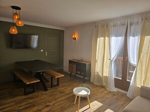 A room with a wooden floor, a dining table with benches, a TV mounted on the wall, a small white stool, and a window with sheer curtains. - 3-room apt, sleeps 6, south-facing balcony (Valloire)