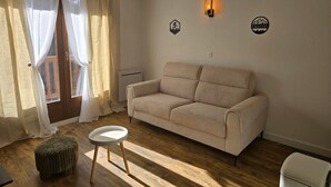 Ein Wohnzimmer mit einem beigen Sofa, einem kleinen runden Tisch und Holzboden. Ein Fenster mit durchsichtigen Vorhängen und eine wandmontierte Leuchte sind ebenfalls vorhanden.