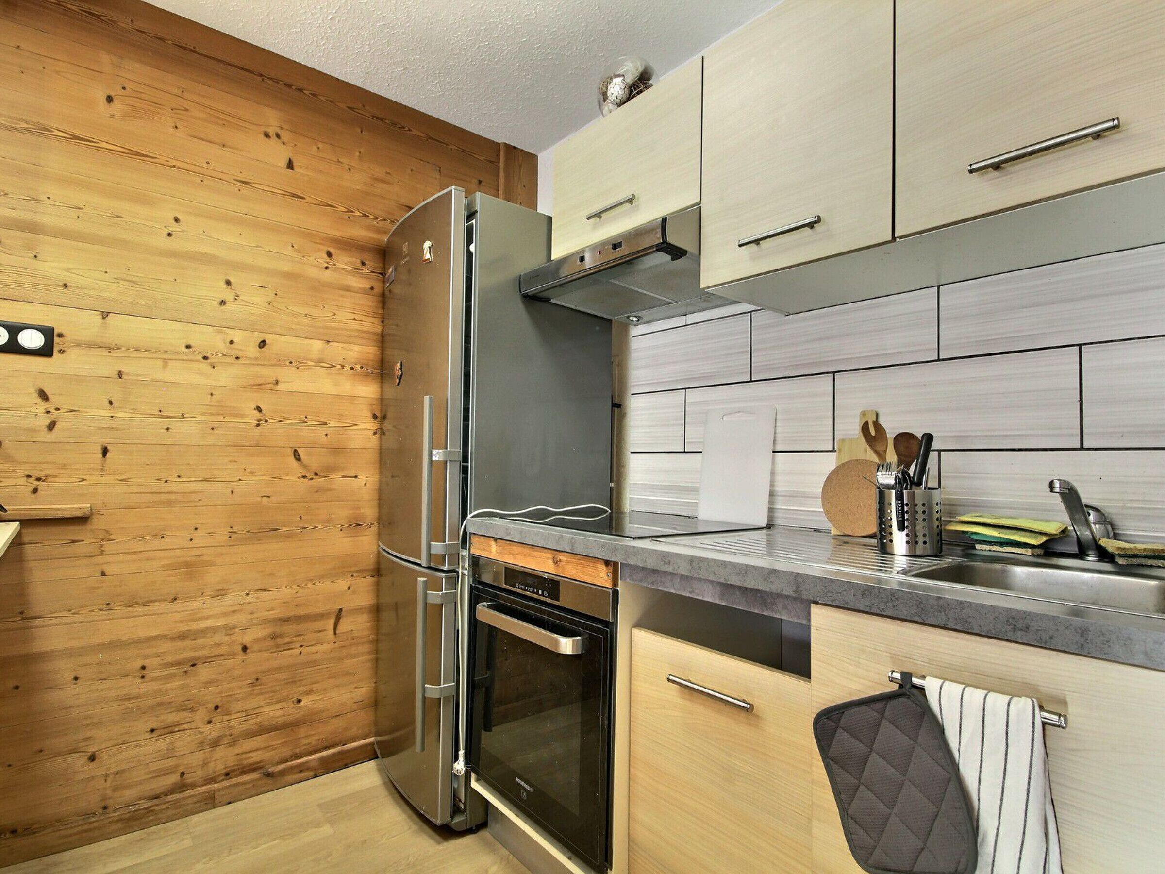 A modern kitchen with wooden walls, a stainless steel refrigerator, an oven, and a countertop with a sink and utensils.