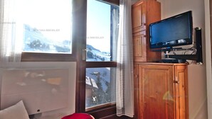Ein Zimmer mit großem Fenster, einem Holzschrank, einem Fernseher und einem roten Sessel.