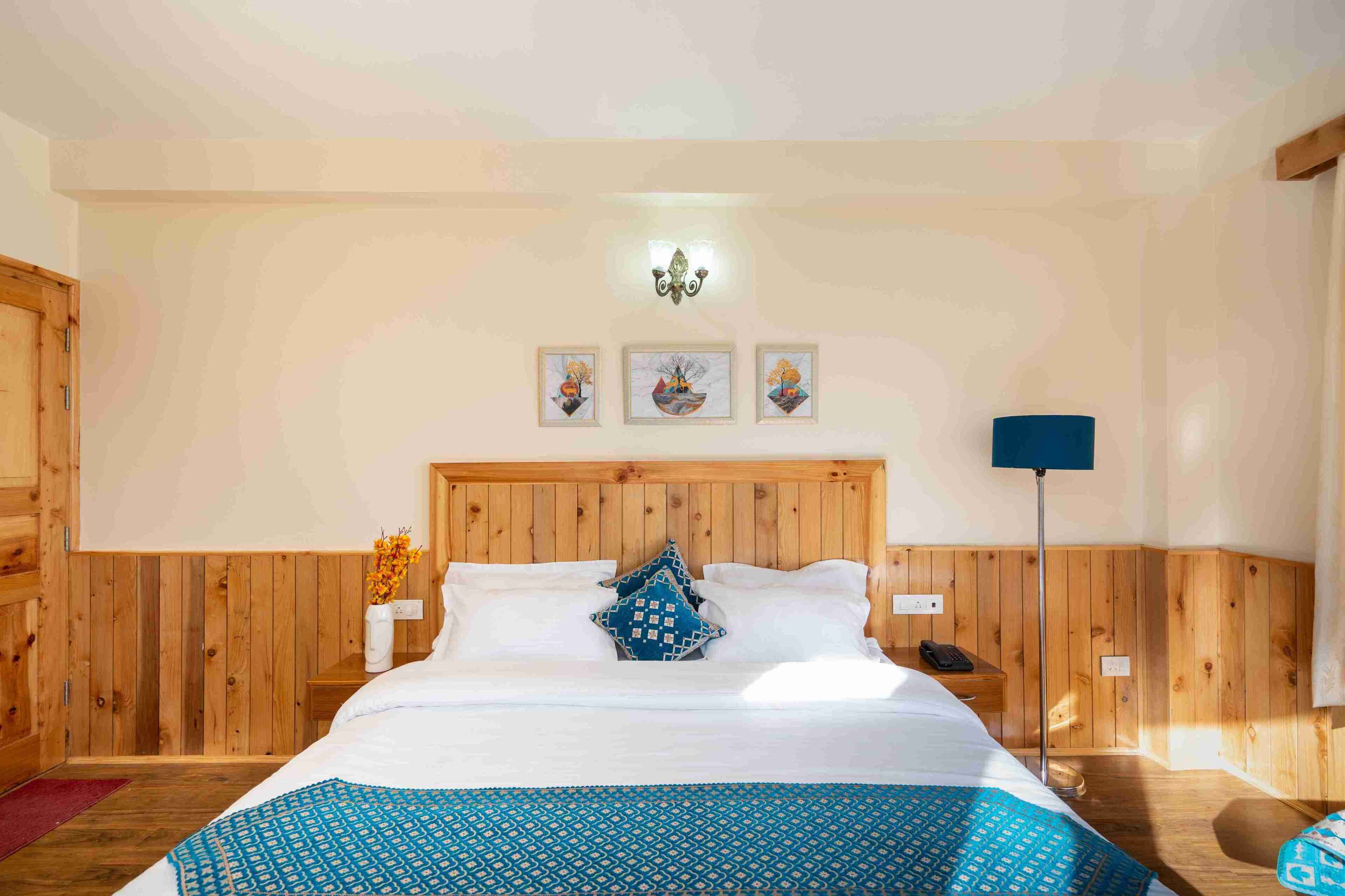 Une chambre à coucher avec une tête de lit en bois, un lit recouvert de draps blancs et d’une courtepointe à motifs bleus, une table de chevet ornée d’un vase de fleurs, et trois tableaux encadrés accrochés au mur.