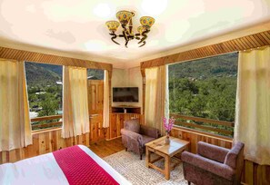 Una habitación con cama, televisión, una mesita, una silla y grandes ventanales con vistas a un paisaje montañoso.