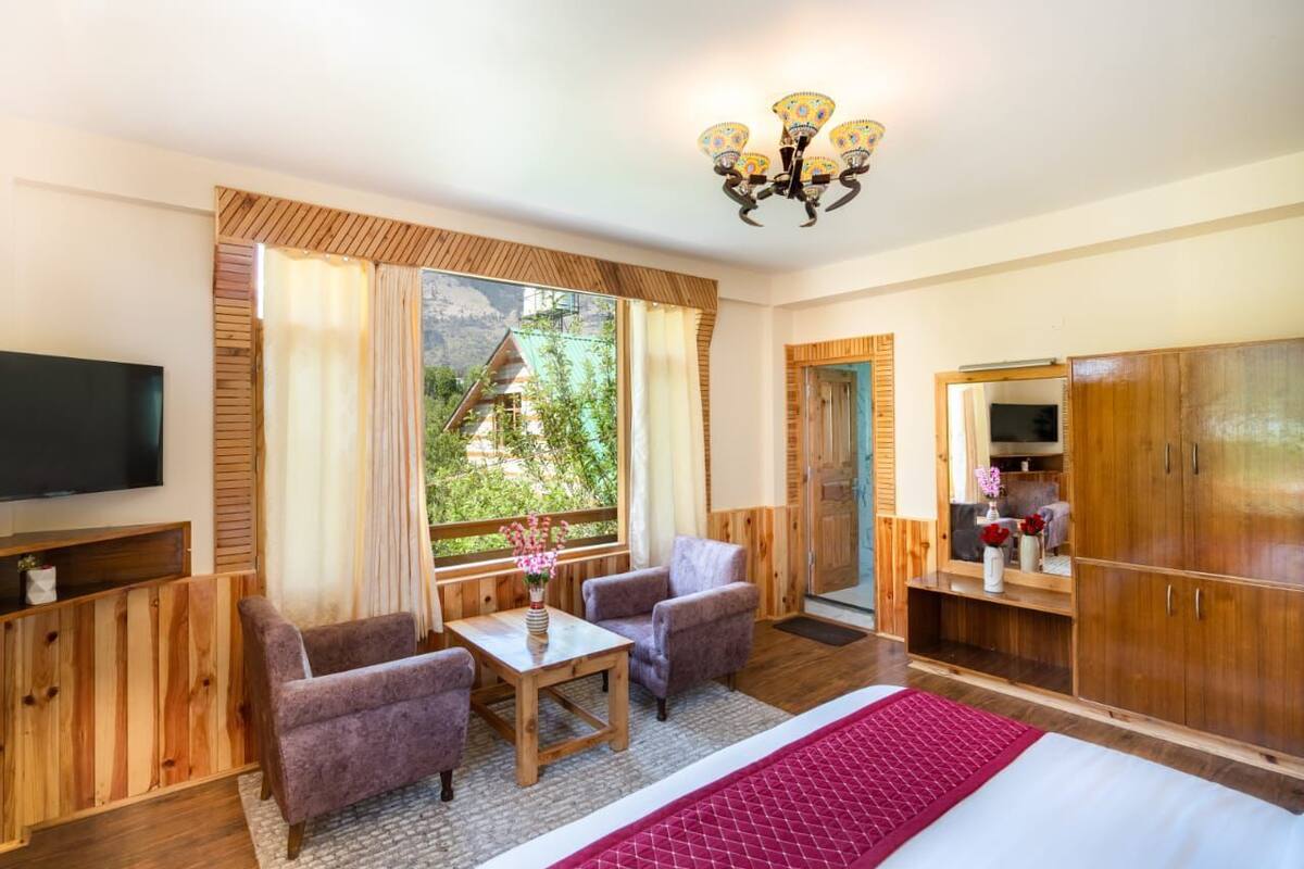 A room with a bed, two armchairs, a small table, a TV, wooden walls, a large window with curtains, and a wooden wardrobe.