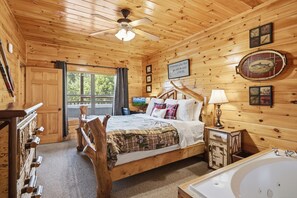 Ein Schlafzimmer einer Holzhütte mit einem Bett, einem Schreibtisch, einem Stuhl, einer Schreibtischlampe, einem wandmontierten Fernseher und einem Fenster mit Vorhängen.