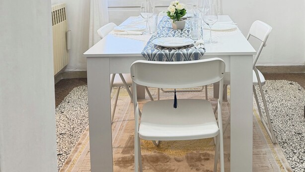 A dining area with a table set for two, white chairs, a chandelier, and a view of buildings through a window with white curtains. - zara'shome (Venezia)
