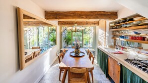 Un espace repas comprenant une longue table en bois, des chaises, un lustre et une vue sur la verdure grâce à de grandes fenêtres.