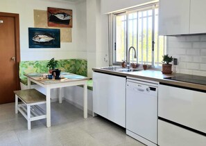A kitchen with a dishwasher, a sink, and a window with a view of trees. There is a table with a chair and a potted plant. - Villa La Yuca: private pool, barbecue area, and private parking (pet-friendly) (Los Alcázares)