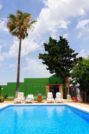 A swimming pool with lounge chairs, a palm tree, and a green building in the background. - Villa La Yuca: private pool, outdoor barbecue, and private parking (pet-friendly) (Los Alcázares)