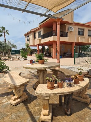 An outdoor seating area with stone tables and chairs, surrounded by potted plants and a building with a balcony. - Villa La Yuca: private pool, barbecue area, and private parking (pet-friendly) (Los Alcázares)