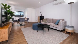 A modern living room with a sofa, ottoman, coffee table, and a TV on a wooden cabinet. There is a dining area with chairs and a table, and a large window with a view of trees. - Casa Susanna (Ponta Delgada)