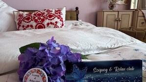 Ein ordentlich gemachtes Bett mit weißer Bettwäsche und einem blumigen Kopfkissen, eine Vase mit lila Blumen und eine auf dem Bett stehende Schachtel mit dem Aufdruck "Szépseg & Relax club".