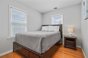 A bedroom with a bed, two windows with blinds, a nightstand with a lamp, and a wooden floor. - Stay just 1 block from the beach & boardwalk in this charming and newly renovated Point Pleasant Beach cottage. Perfect for families and small groups, this cozy beach retreat combines an unbeatable location with comfortable indoor and outdoor spaces (Point Pleasant Beach)