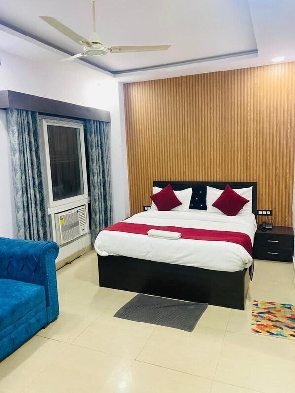 A neatly arranged bedroom with a bed, a blue armchair, a ceiling fan, and a wooden panel wall. - Hotel Paras R Inn (Lucknow)