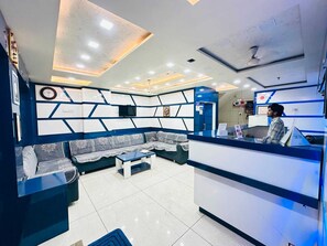 A reception area with a sofa, a reception desk, and a wall-mounted television. - Hotel Hari Palace (Rajkot)