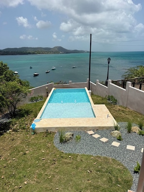 A rectangular pool with a clear blue water surface, surrounded by a low stone wall, with a view of the sea and boats in the background. - Frente Del Mar  Oceanfront Pool Villa  Walk to Ferry, Dining & Lighthouse (Vieques)