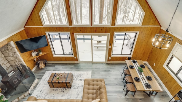 A spacious loft with a high ceiling, wooden walls, a stone fireplace, a flat-screen TV, a dining area with a long table, and large windows offering a view of the outdoors. - Chalet-view Home w/ Fire Pit & Deck in Clifton Twp (Gouldsboro)