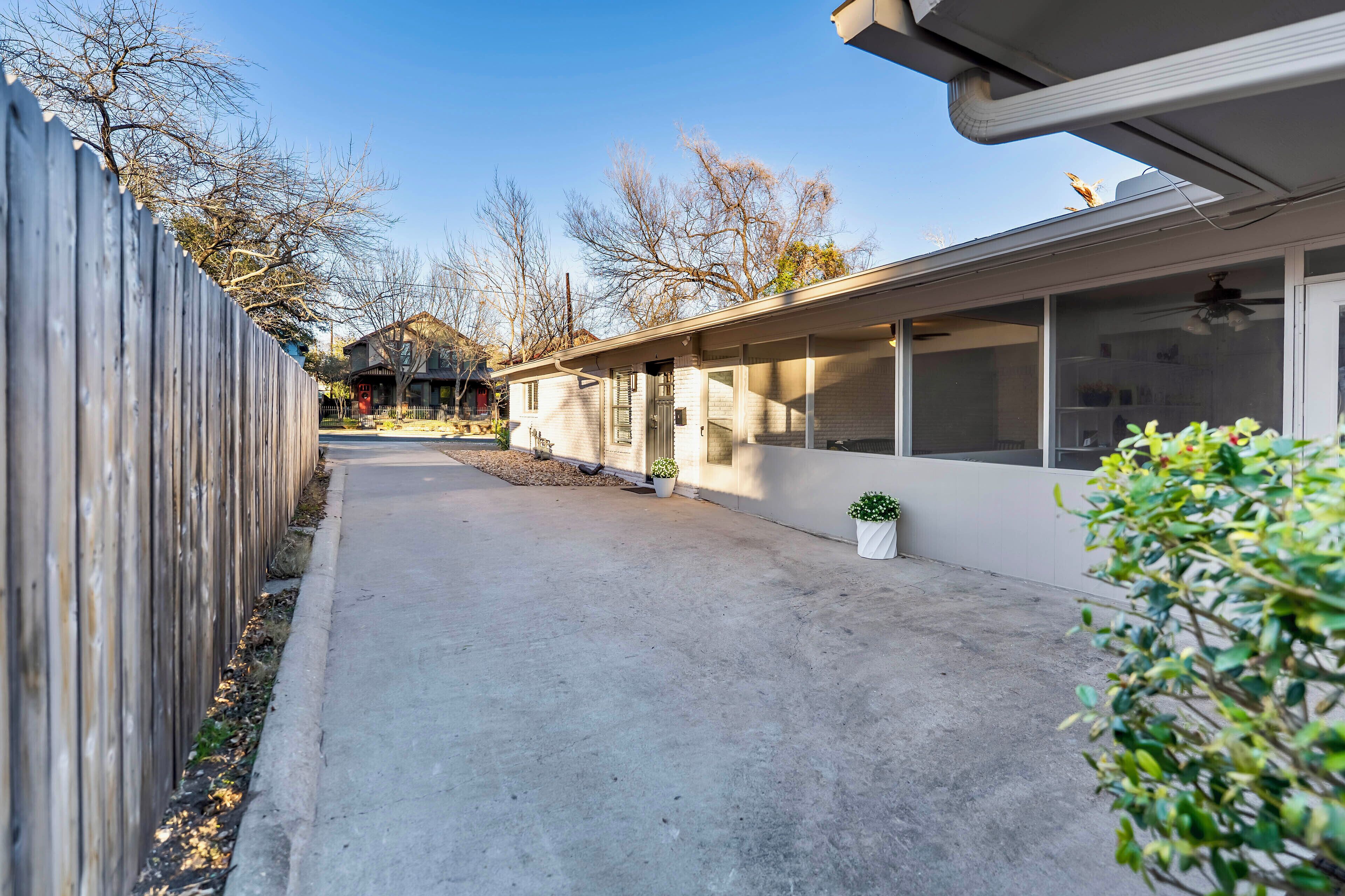 A residential driveway with a wooden fence, potted plants, and a house with large windows and a covered porch.