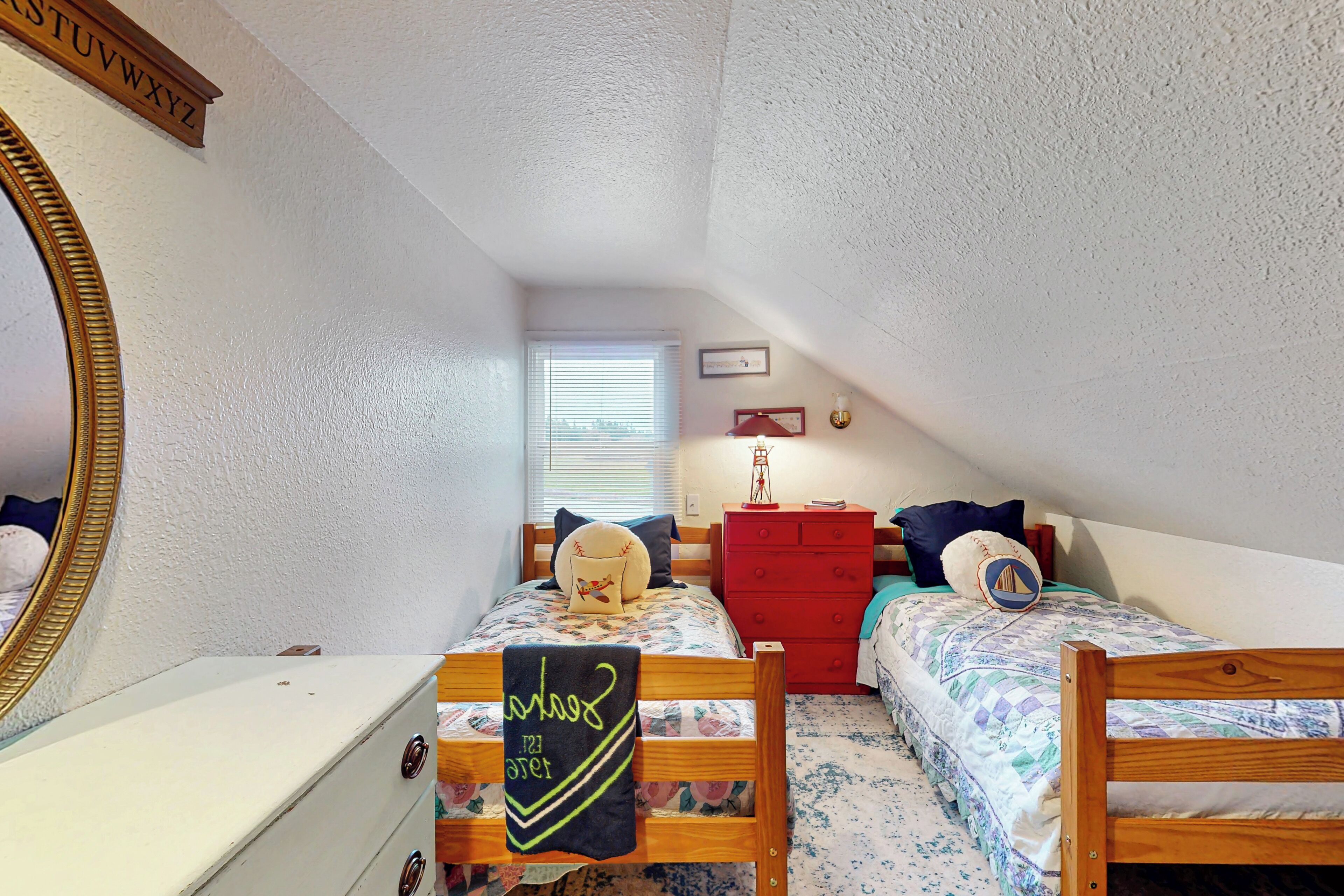Un dormitorio pequeño y luminoso con dos camas de madera, un aparador rojo, un espejo y una ventana con persianas.