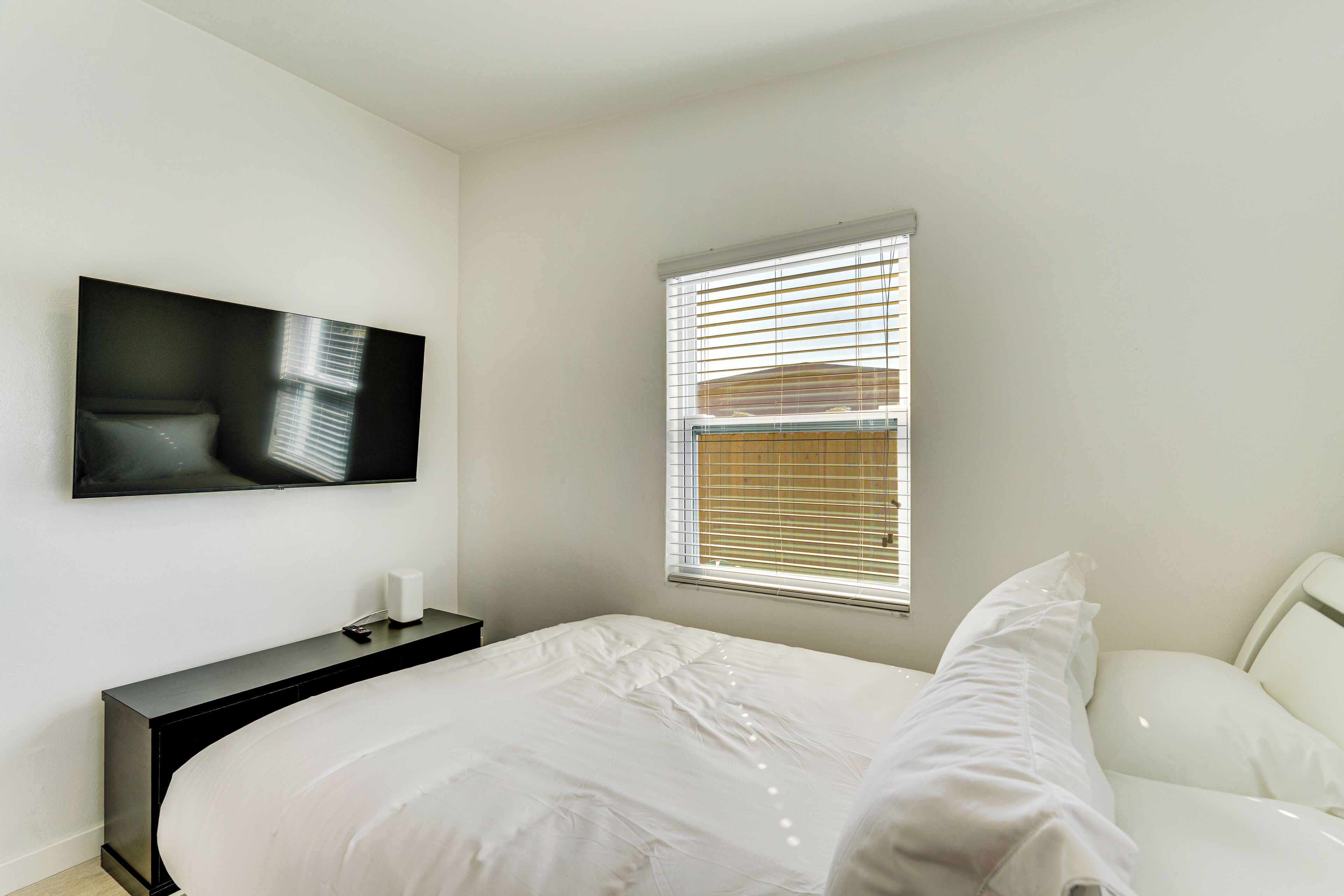 A modern bedroom with a large bed, a bedside table, a TV mounted on the wall, and a window with blinds.