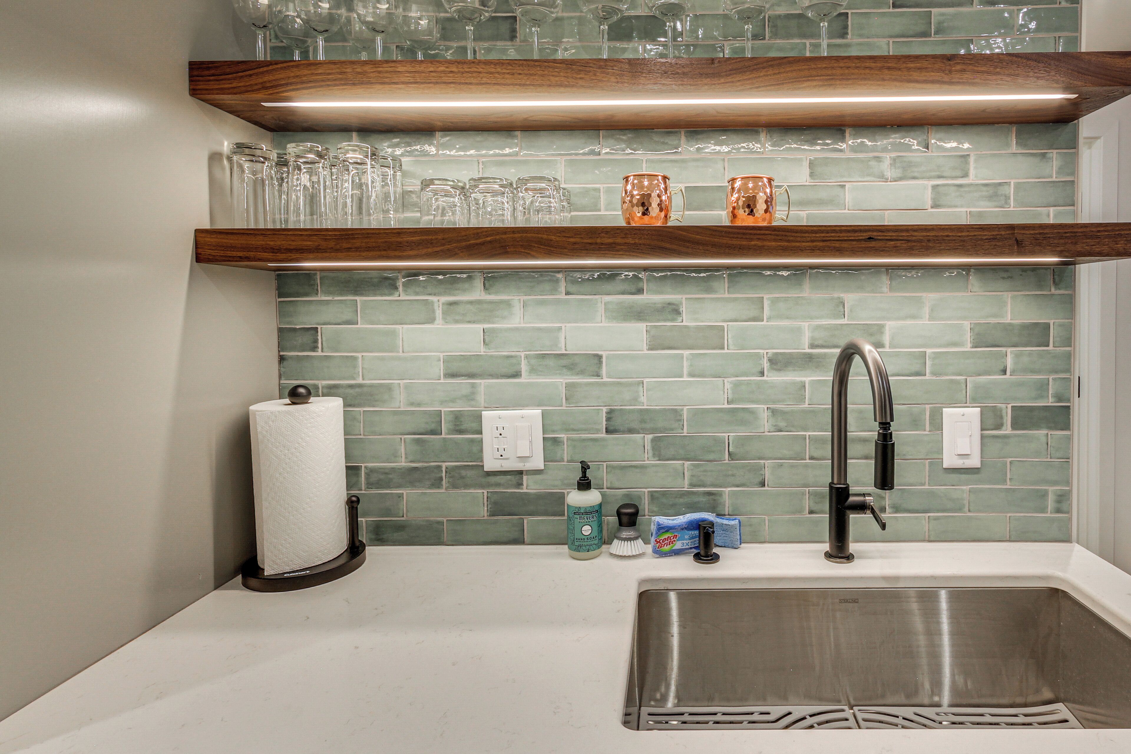 A kitchen with a stainless steel sink, a wooden shelf above holding glassware, and a green-tiled backsplash.