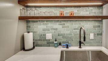 A kitchen with a stainless steel sink, a wooden shelf above holding glassware, and a green-tiled backsplash.
