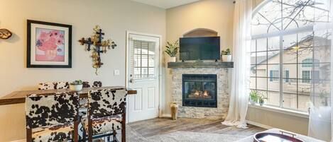 A living room with a fireplace, a dining area with a table and chairs, and a large window with a view of buildings.