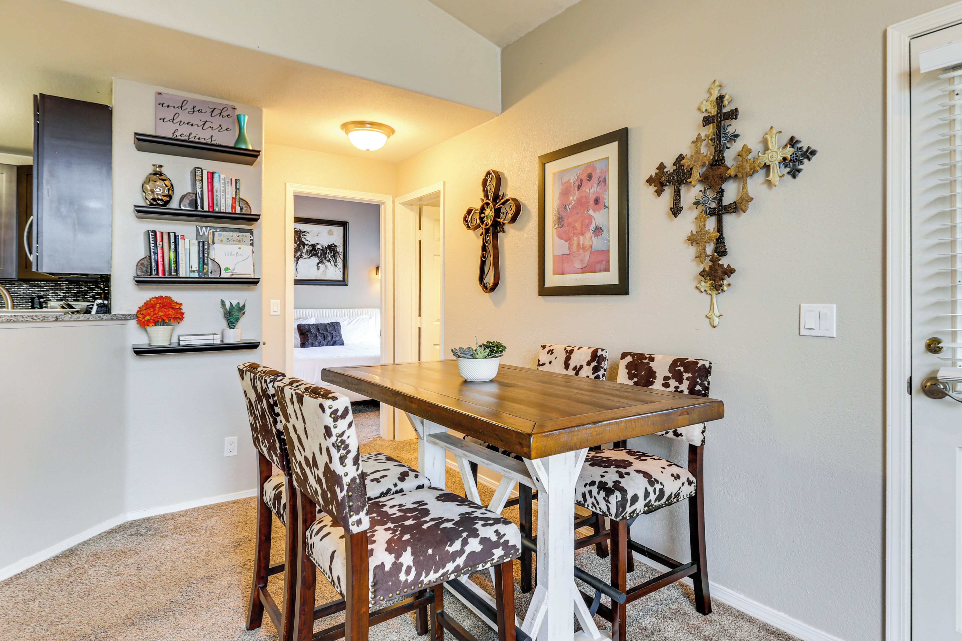 Un coin repas avec une table en bois et des chaises recouvertes de cuir, une bibliothèque remplie de livres et d’objets décoratifs, et un mur orné d’une croix et d’un tableau encadré.