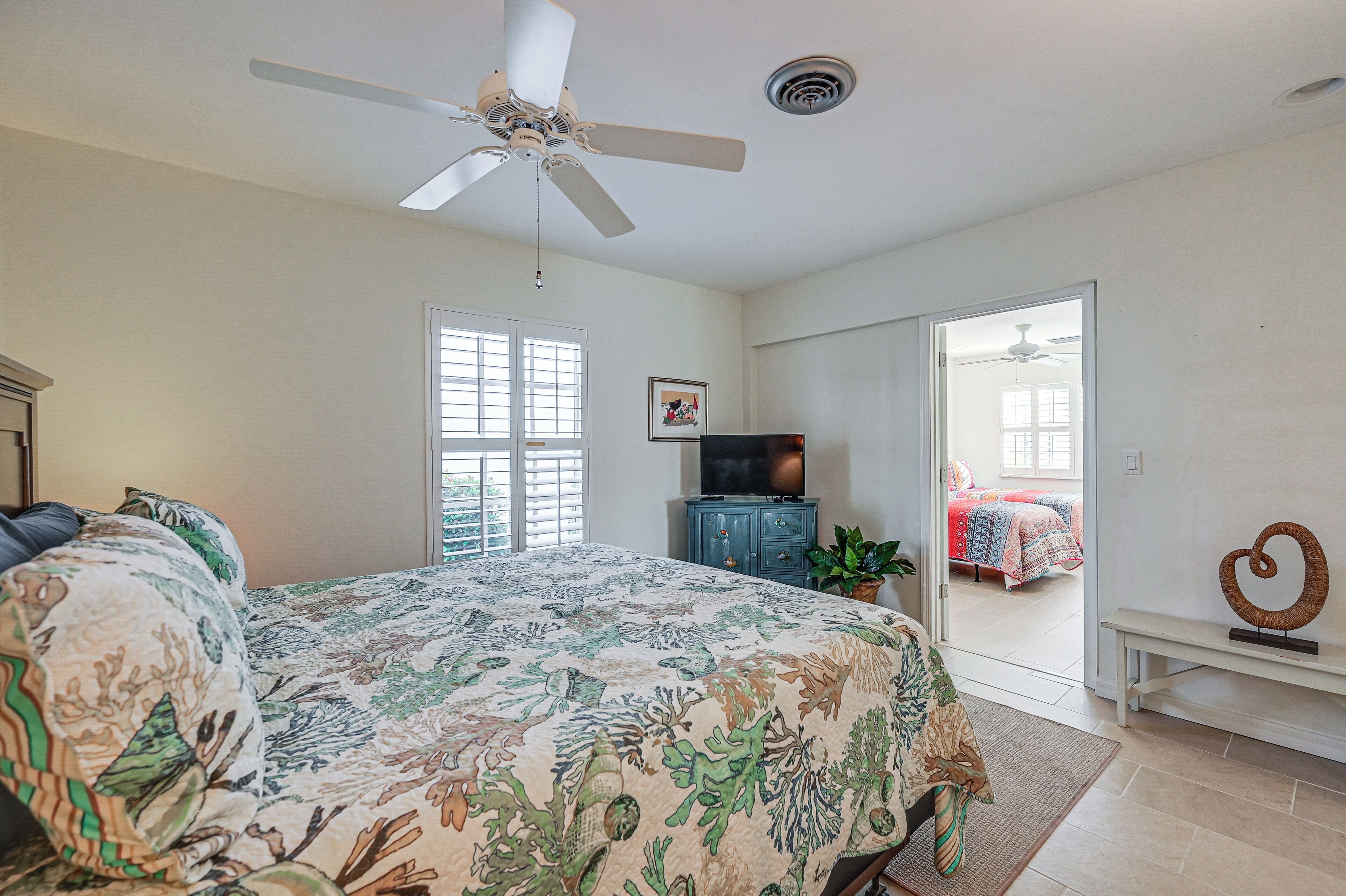 A bedroom with a bed, a ceiling fan, a TV, and a door leading to another room.
