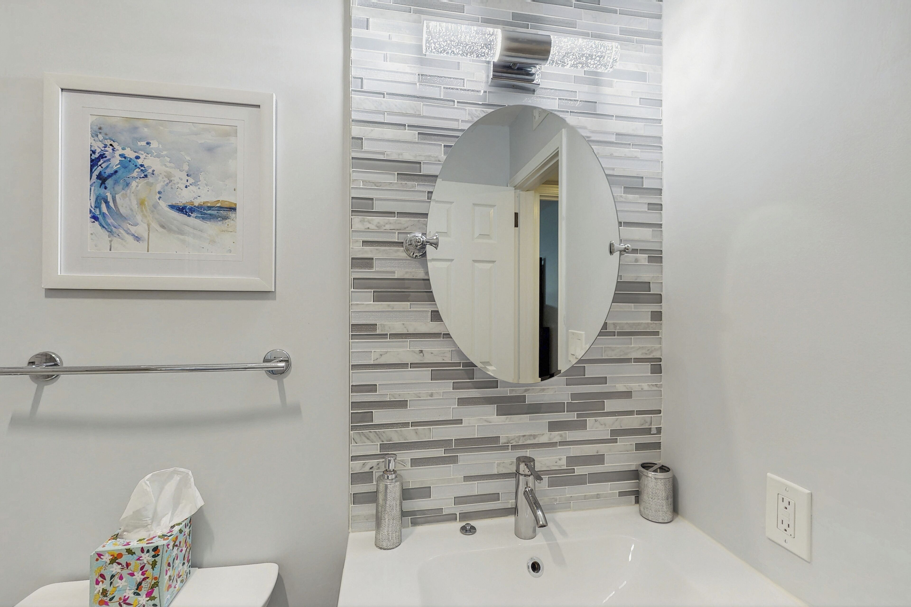 A bathroom with a tiled backsplash, a framed artwork, a round mirror, a toilet, and a sink with a faucet.