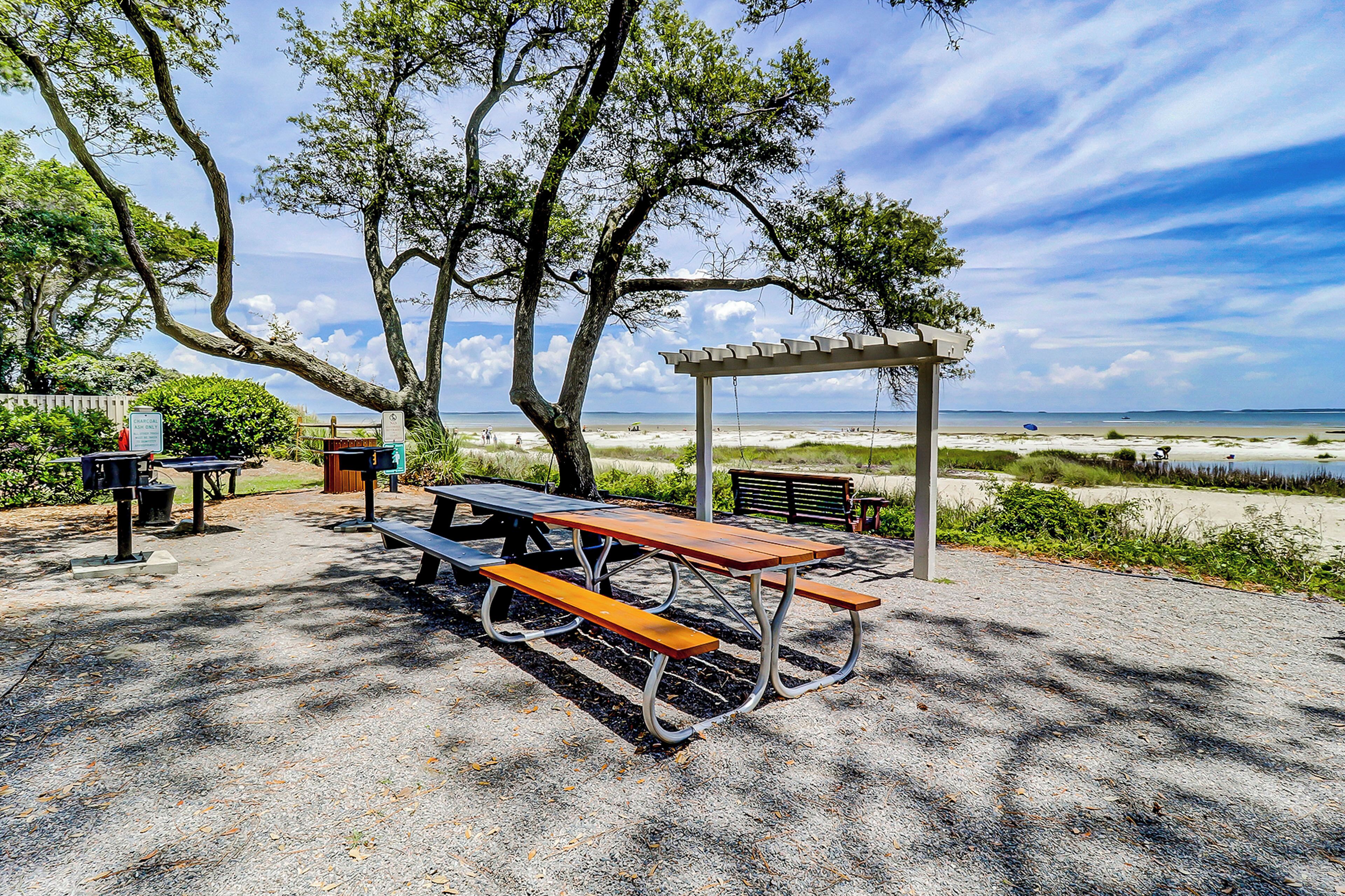 A picnic area with tables and benches, surrounded by trees and a view of the beach.