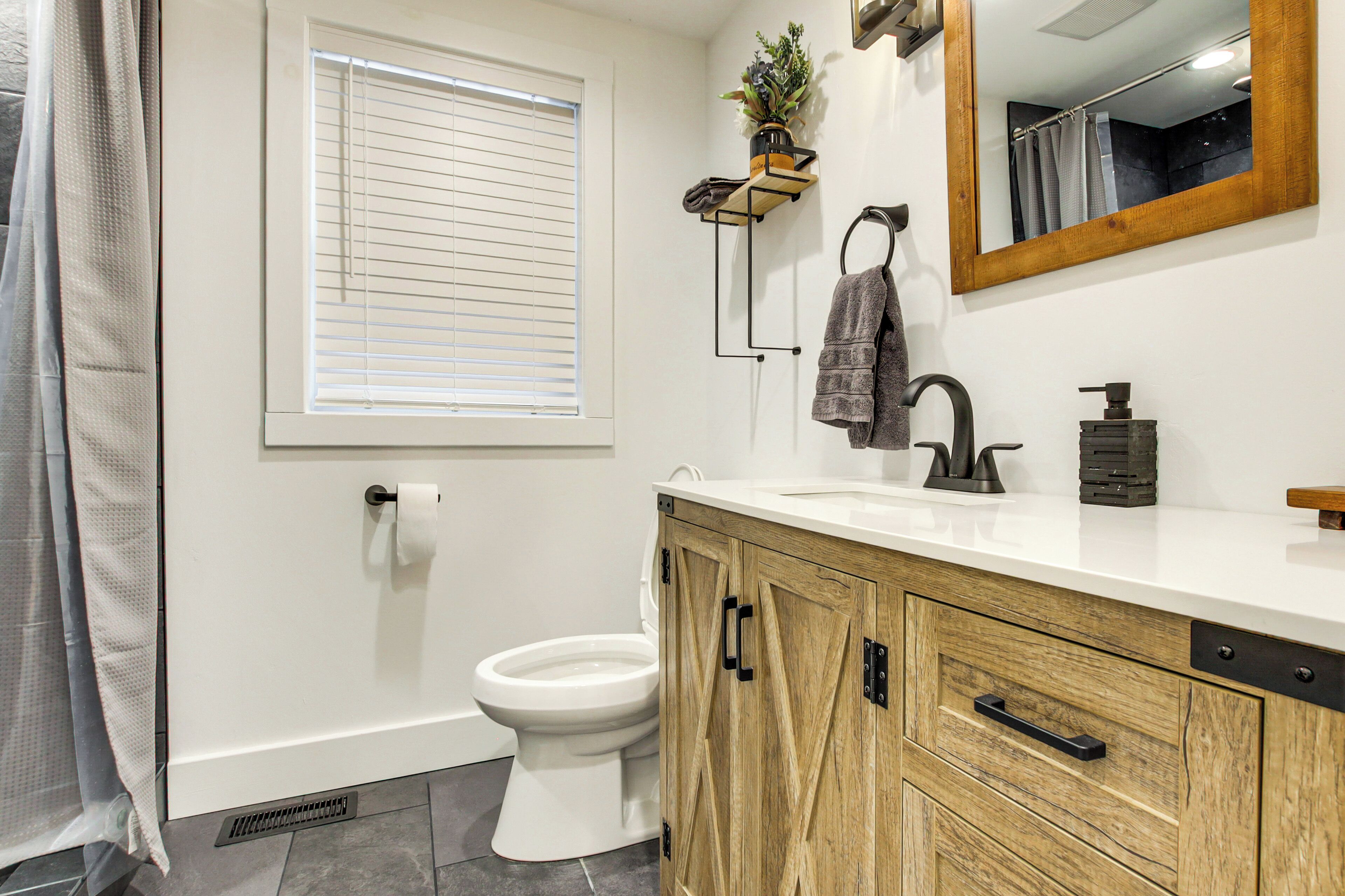 A bathroom with a toilet, sink, wooden cabinet, mirror, and a window with blinds.