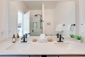 A modern bathroom with two sinks, a large mirror, a shower, and a potted plant.