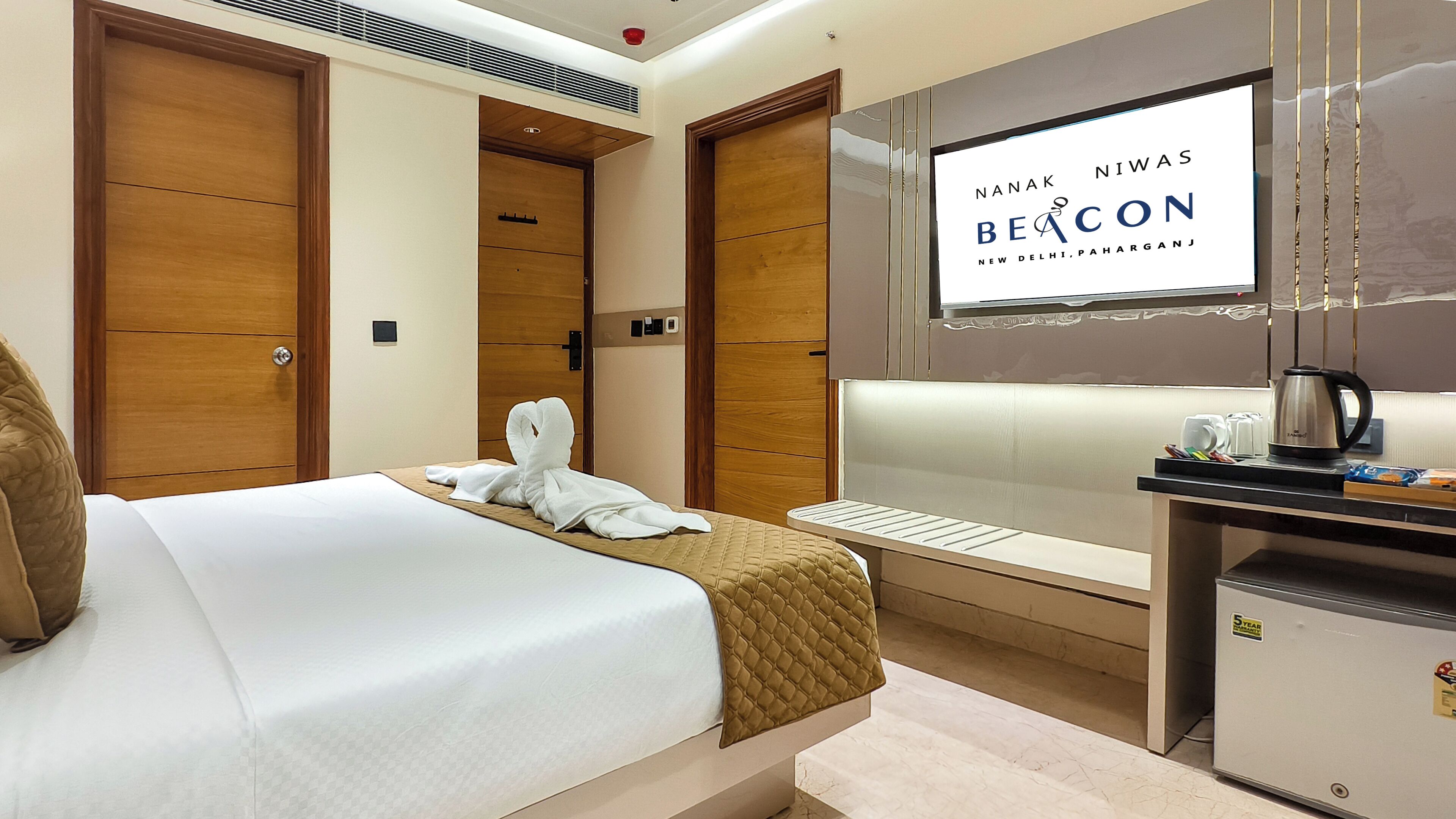 A hotel room with a bed, a desk with a kettle, and a TV displaying 'Nanak Niwas Beacon'.