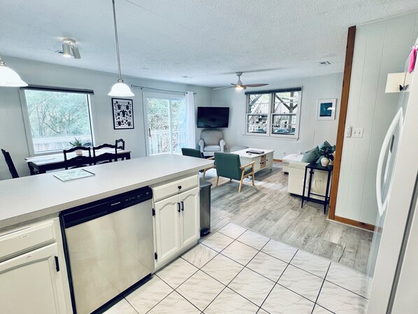 A modern kitchen with a dishwasher, a dining area with chairs, a TV, and a ceiling fan. - Pets, Pool & Spa - Tranquil Treetop Family Home (Kitty Hawk)