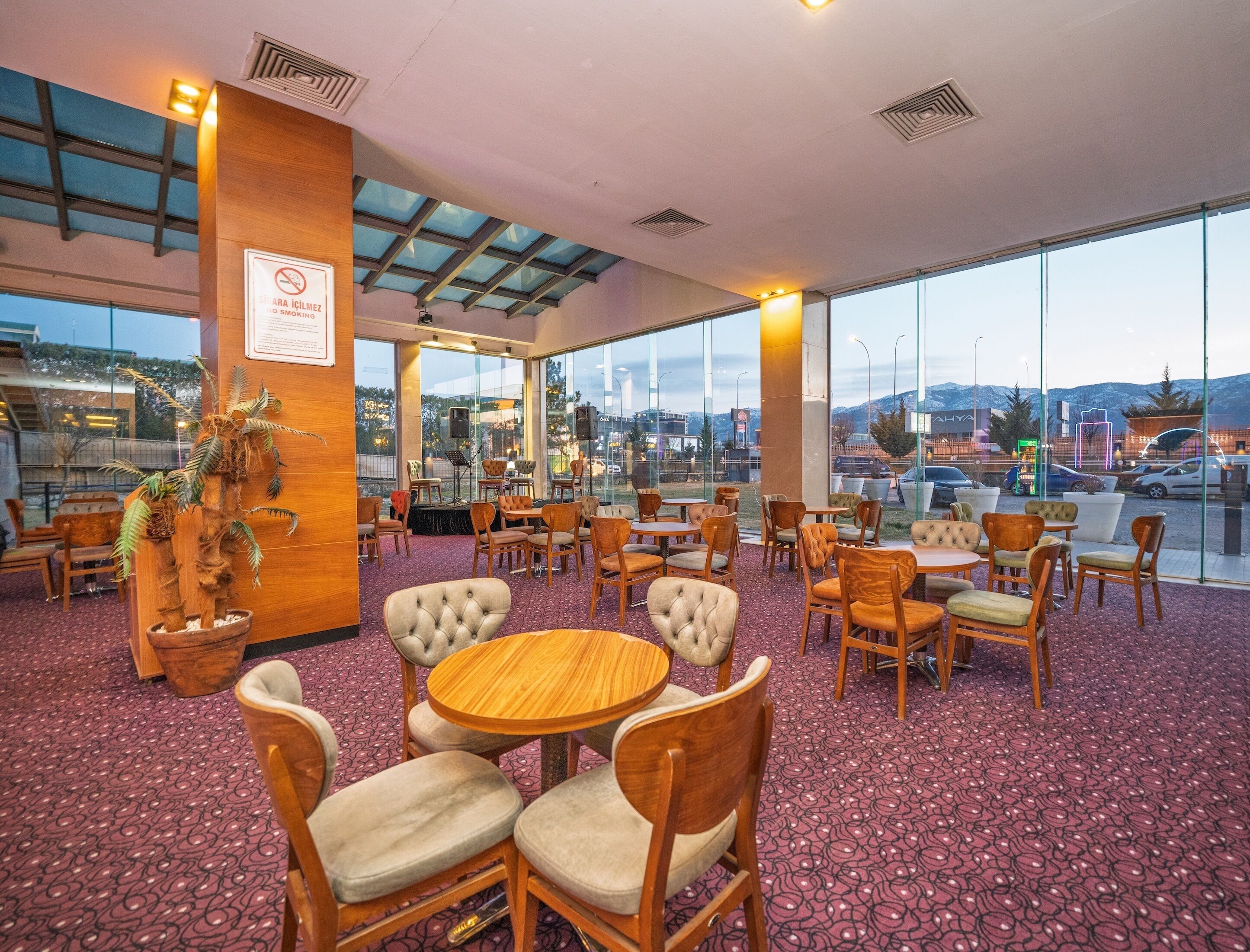 A spacious indoor seating area with multiple tables and chairs, large windows offering a view of the cityscape, and a decorative plant arrangement.