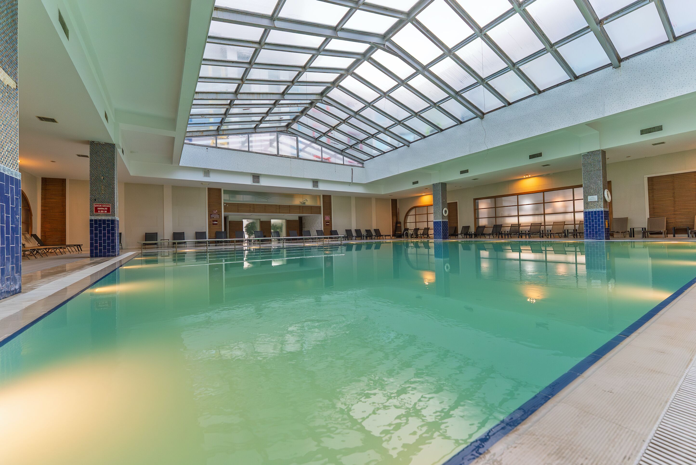 An indoor swimming pool with a glass ceiling, multiple lanes, and a seating area with chairs.