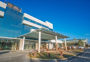 A modern hotel building with a glass facade, a covered entrance, and a landscaped area with shrubs and a small pond.