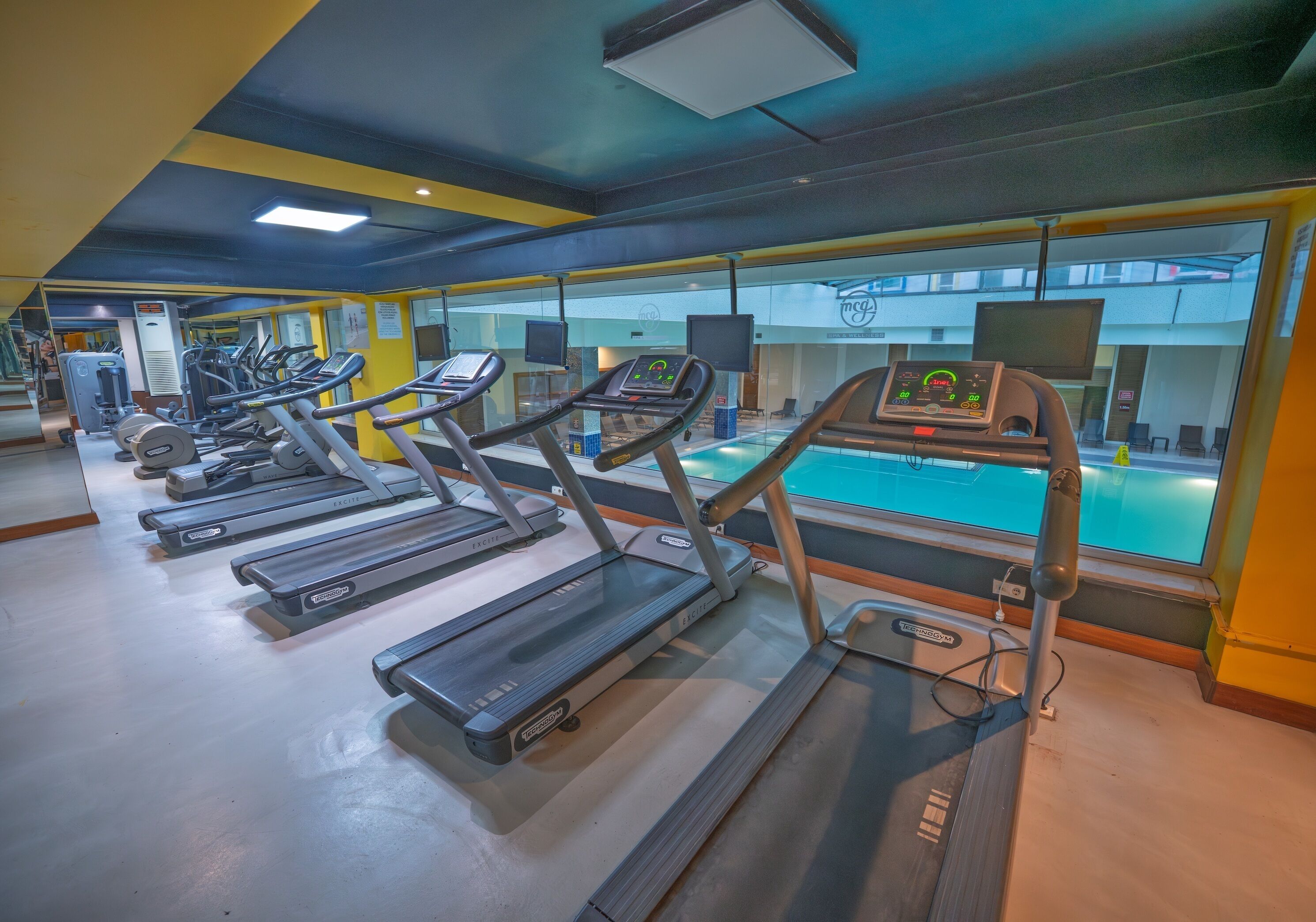 Un gimnasio con varias cintas de correr, equipamiento de ejercicio y una zona de piscina visible a través de las ventanas.