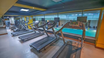 Un gimnasio con varias cintas de correr, equipamiento de ejercicio y una zona de piscina visible a través de las ventanas.
