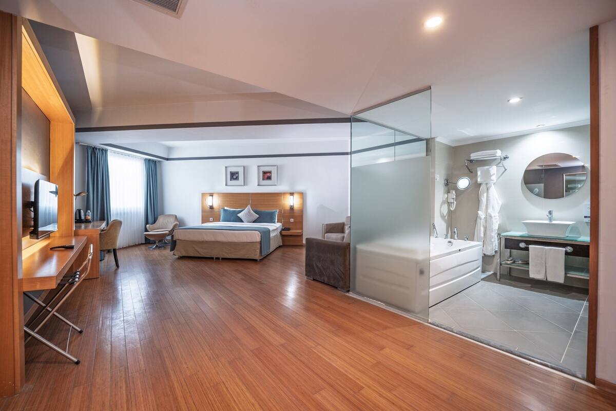 Una habitación de hotel moderna con piso de madera, una cama con cabecera, un área de descanso con escritorio y sillas, una pared de vidrio y un baño con bañera y lavamanos.
