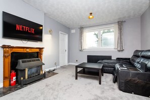 A living room with a fireplace, a leather sofa, a TV mounted on the wall, and a window with curtains. - 4 Bedroom House - Free Parking (Aberdeen)