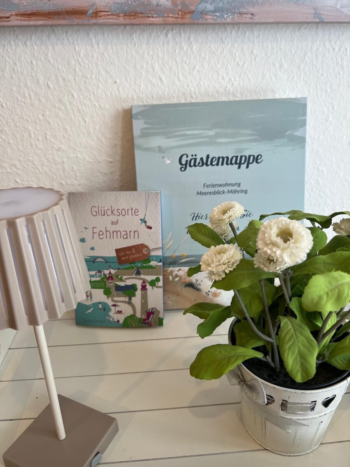 Une pièce avec une lampe blanche, un livre intitulé « Glücksorte aus Fehmarn » et une carte encadrée portant la mention « G ästemappe ».