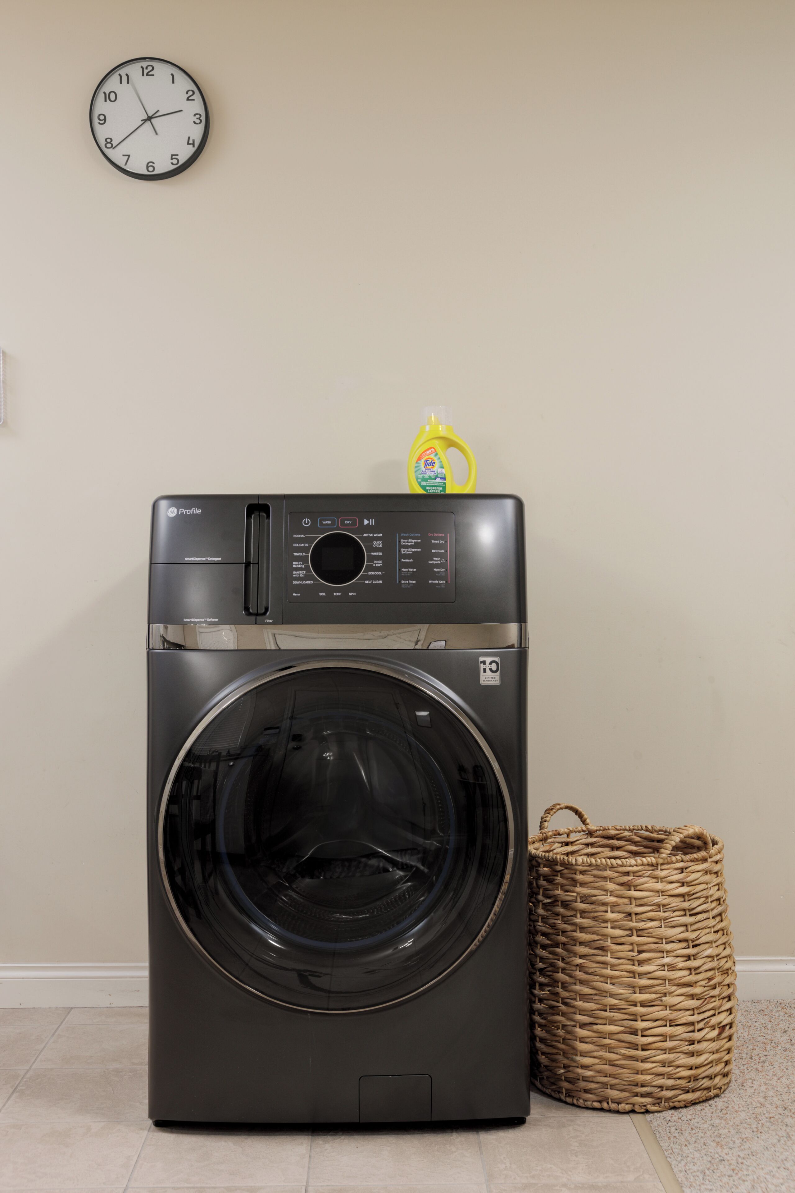 A front-loading washing machine with a control panel, a clock on the wall, and a wicker basket.
