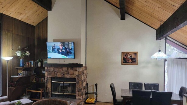 Een gezellige woonkamer met een stenen openhaard, een aan de muur bevestigde televisie, een eethoek met stoelen en een boekenplank vol boeken en decoratieve objecten.