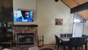 Een woonkamer met een stenen open haard, een aan de muur bevestigde televisie, een eettafel met stoelen, en een ingelijst schilderij.