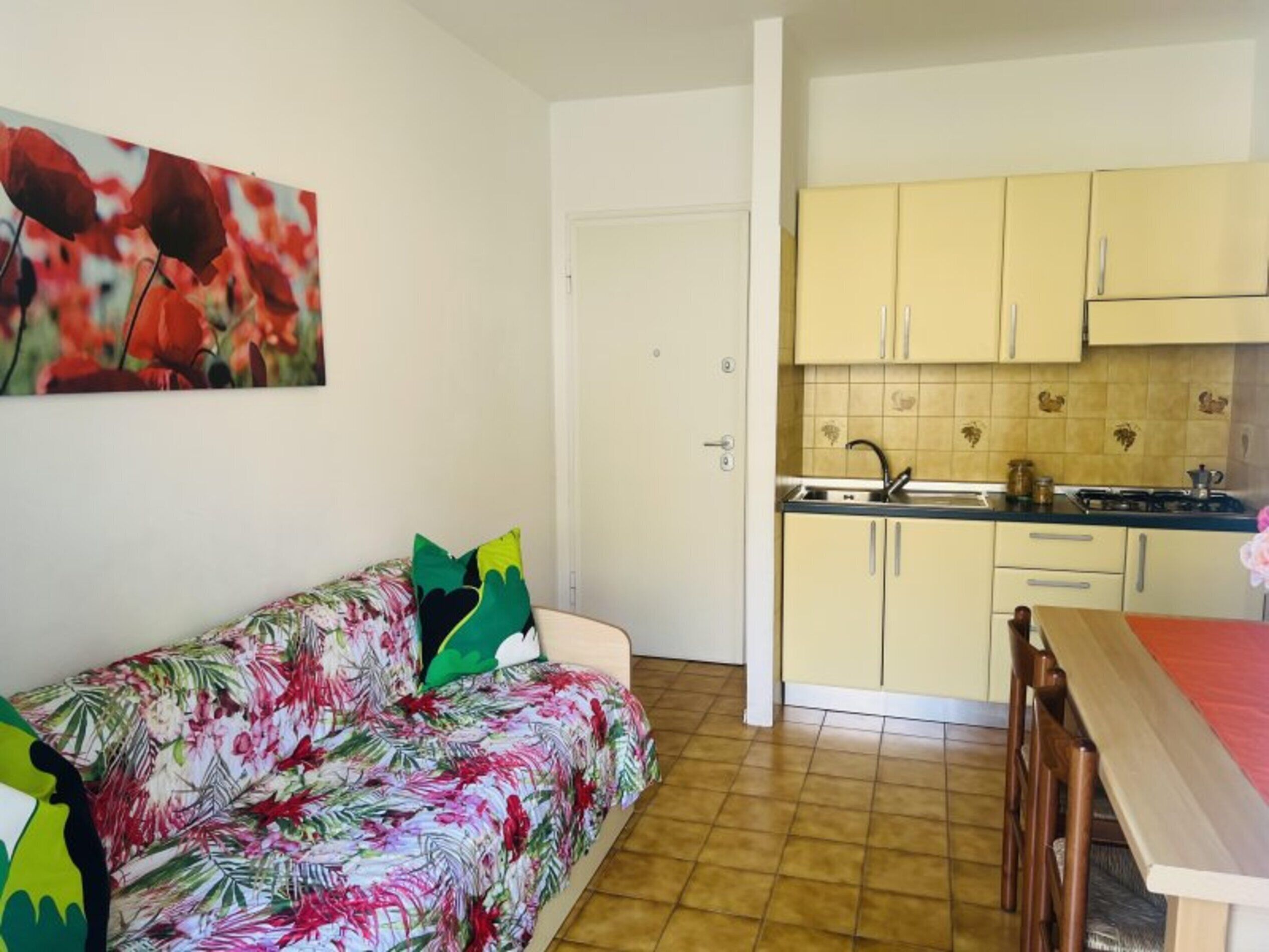 Ett kompakt bostadsutrymme med ett litet kök, en soffa med blommigt tyg och en dörr som leder till ett annat rum.