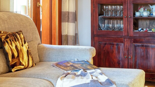 Ein Wohnzimmer mit einer Couch, einer Zeitschrift, einer gemusterten Decke und einem Holzschrank mit Glastüren, der Gläser beherbergt.