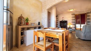Ein gemütlicher Wohnbereich mit einem Holzesstisch, Stühlen und einer Couch. Es gibt eine Küchenzeile mit einem Kühlschrank, einem Holzschrank und einer kleinen Kochinsel mit einer Schüssel und Pflanzen. Der Raum hat eine Backsteinwand und einen gefliesten Boden.