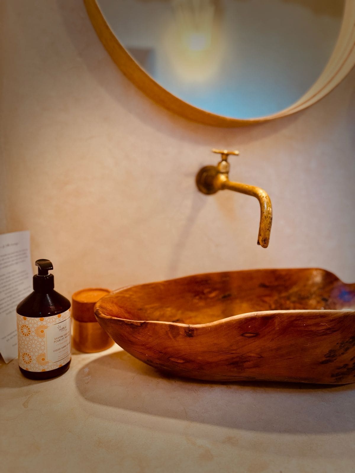 A wooden basin with a gold faucet, a bottle of liquid soap, and a candle on a bathroom counter.