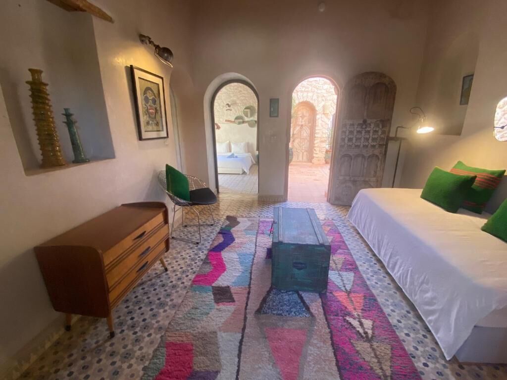 Une chambre à coucher avec un lit, une commode, une chaise et un tapis coloré. Deux arcades mènent à une autre pièce.